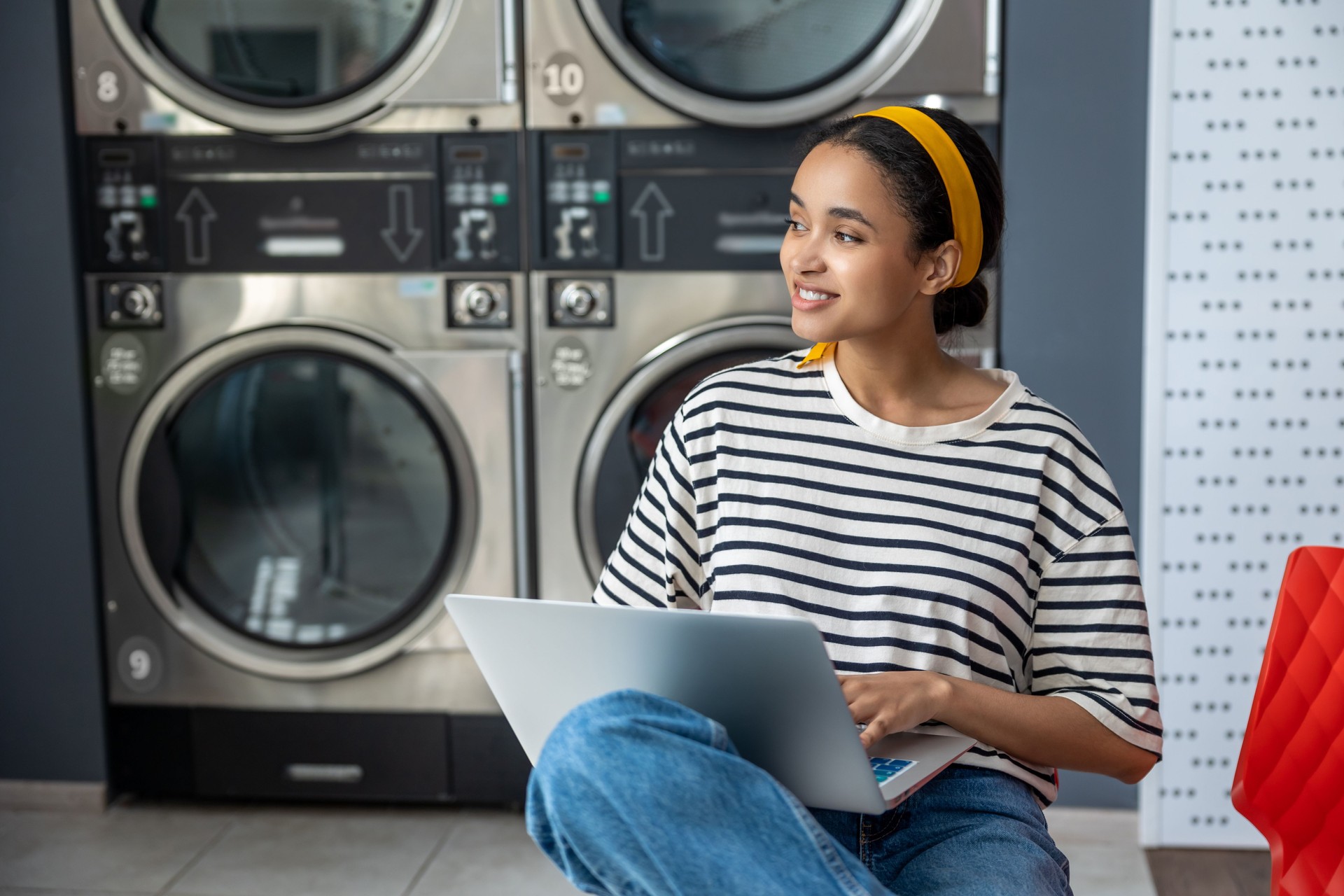 Woman sitting near washing machine using laptop while working remotely on online task
